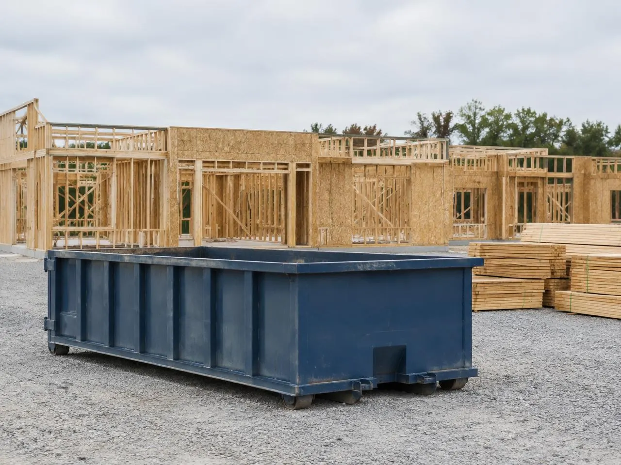 Empty navy roll-off dumpster on a gravel construction site beside a wood-framed building and stacked lumber